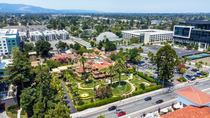 Aerial view of historic Winchester Mystery House with its lush gardens, surrounded by modern office and residential buildings and a busy street - San Jose, California, USA - July 14, 2024